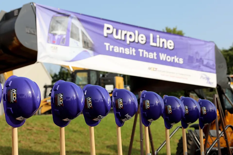 Line of purple construction helmets on shovels in front of Purple Line banner for photo op at BRT line groundbreaking.